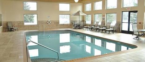 Una piscina interior con suelo de baldosas, ventanas iluminadas por el sol y sillones de descanso.