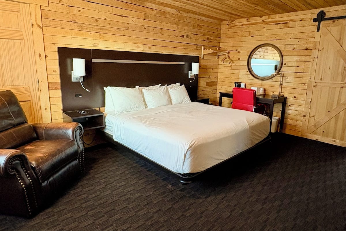 A wooden-paneled room with a bed, a leather chair, a bedside table, and a mirror on the wall.