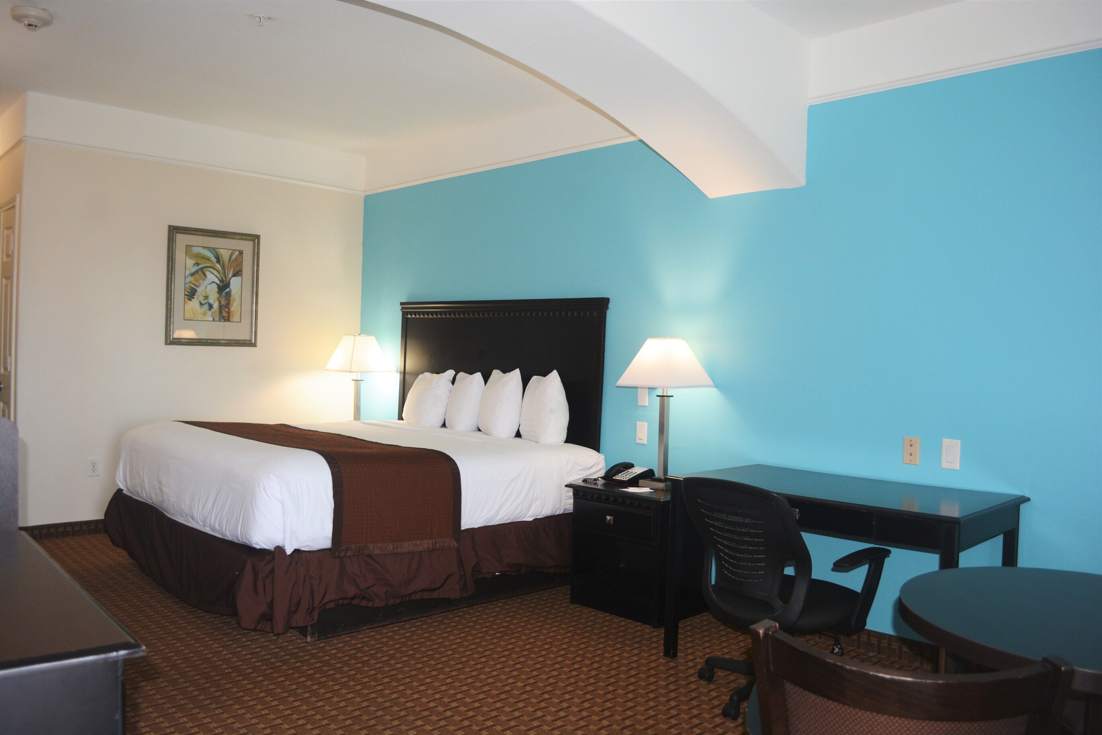 A hotel room with a bed, desk, and chair, featuring a blue accent wall and a patterned carpet.