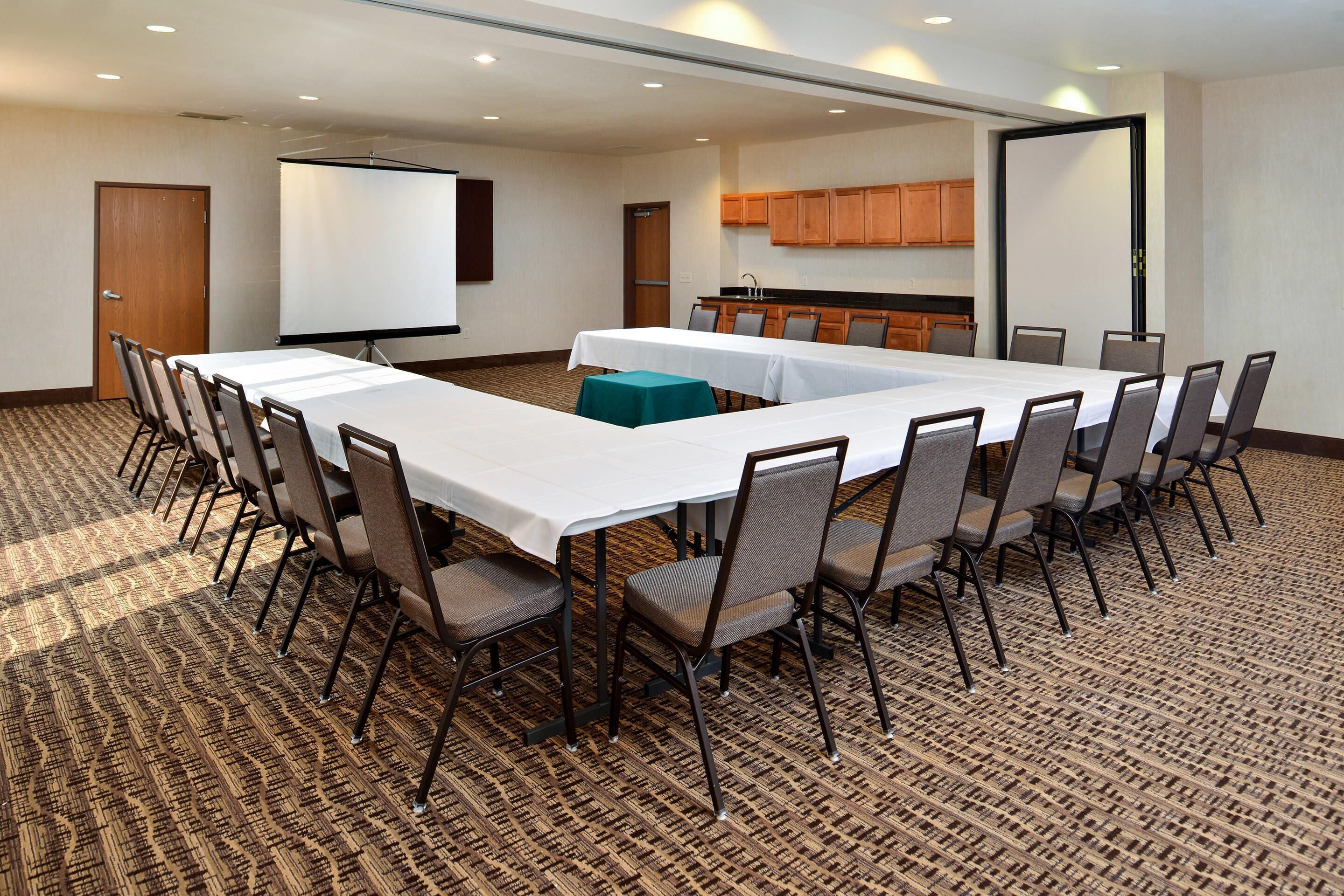 A conference room with a long table, chairs, a projector screen, and a kitchenette.