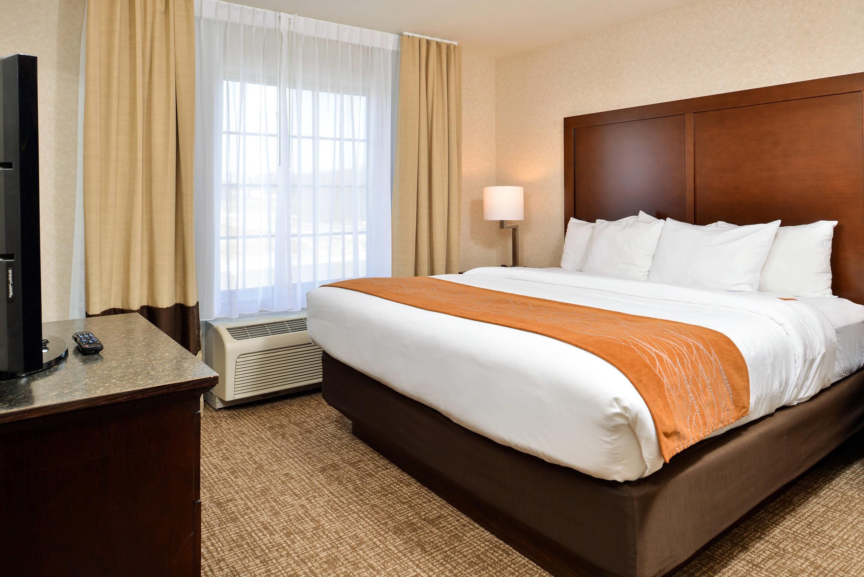 A hotel room with a bed, a desk with a TV and remote, a lamp, and an air conditioning unit.