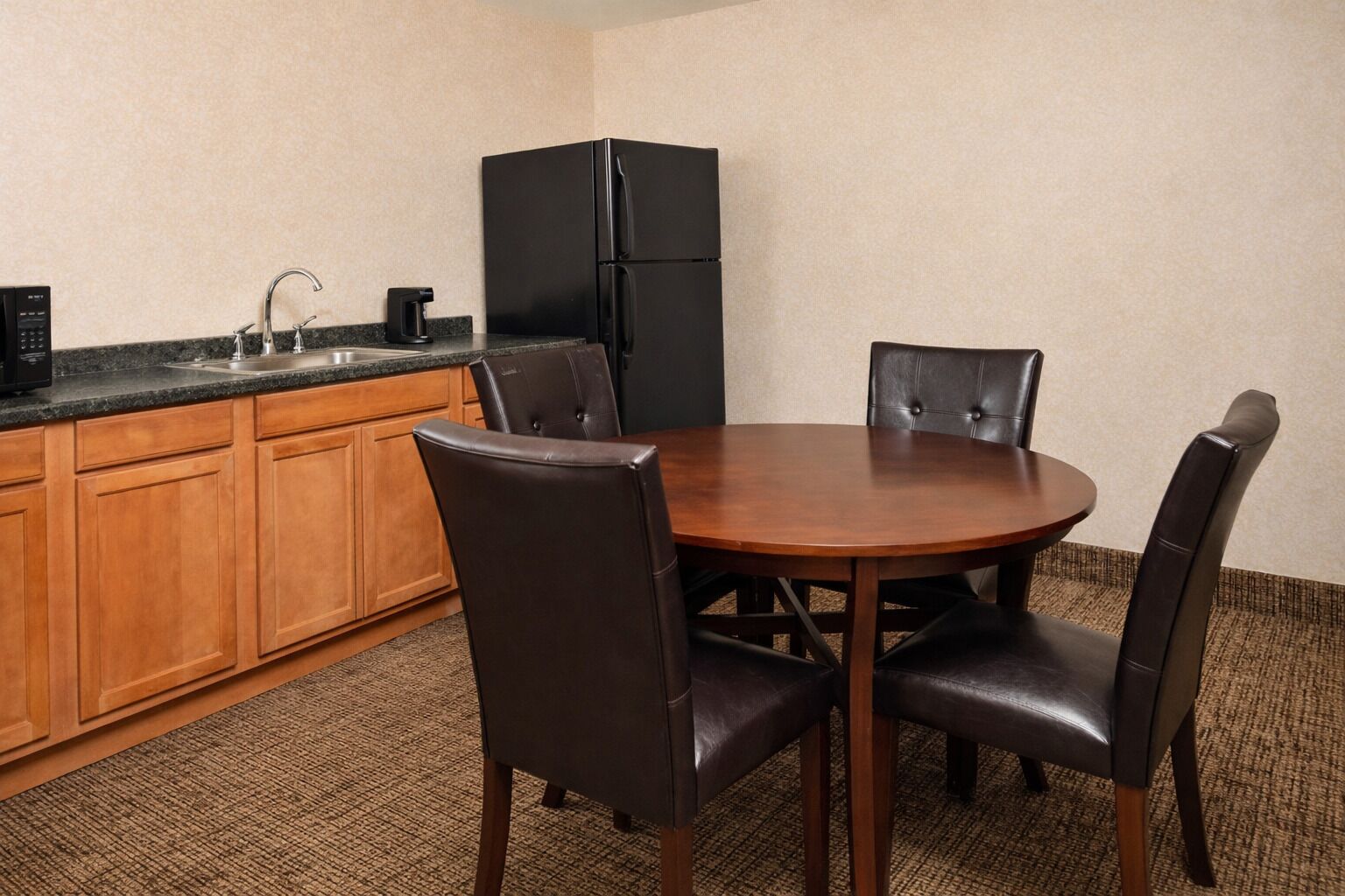 A compact kitchen with a black refrigerator, wooden cabinets, a sink, and a dining table with four chairs.