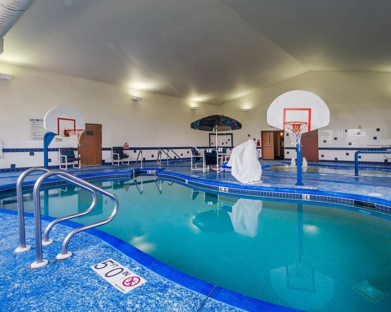 An indoor pool with two basketball hoops, lounge chairs, and a covered area.