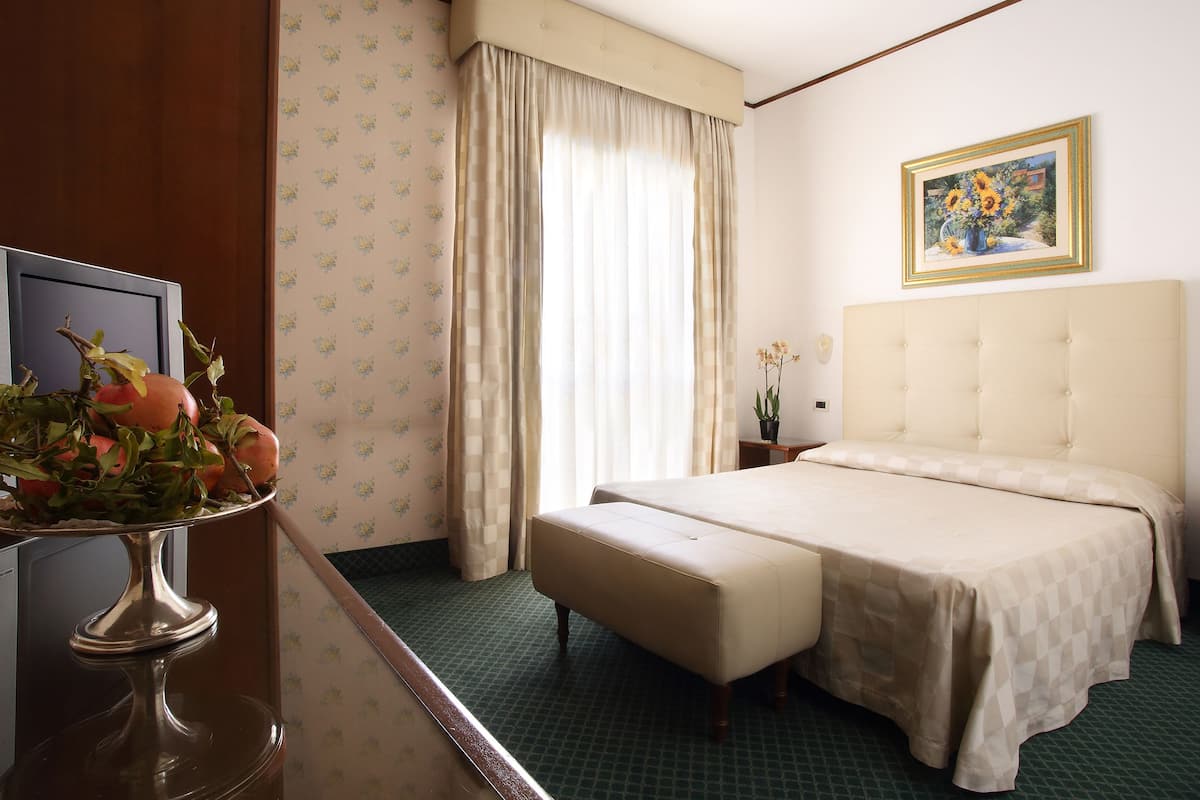 A hotel room with a bed, a small table with a vase of flowers, a painting on the wall, and a window with curtains.