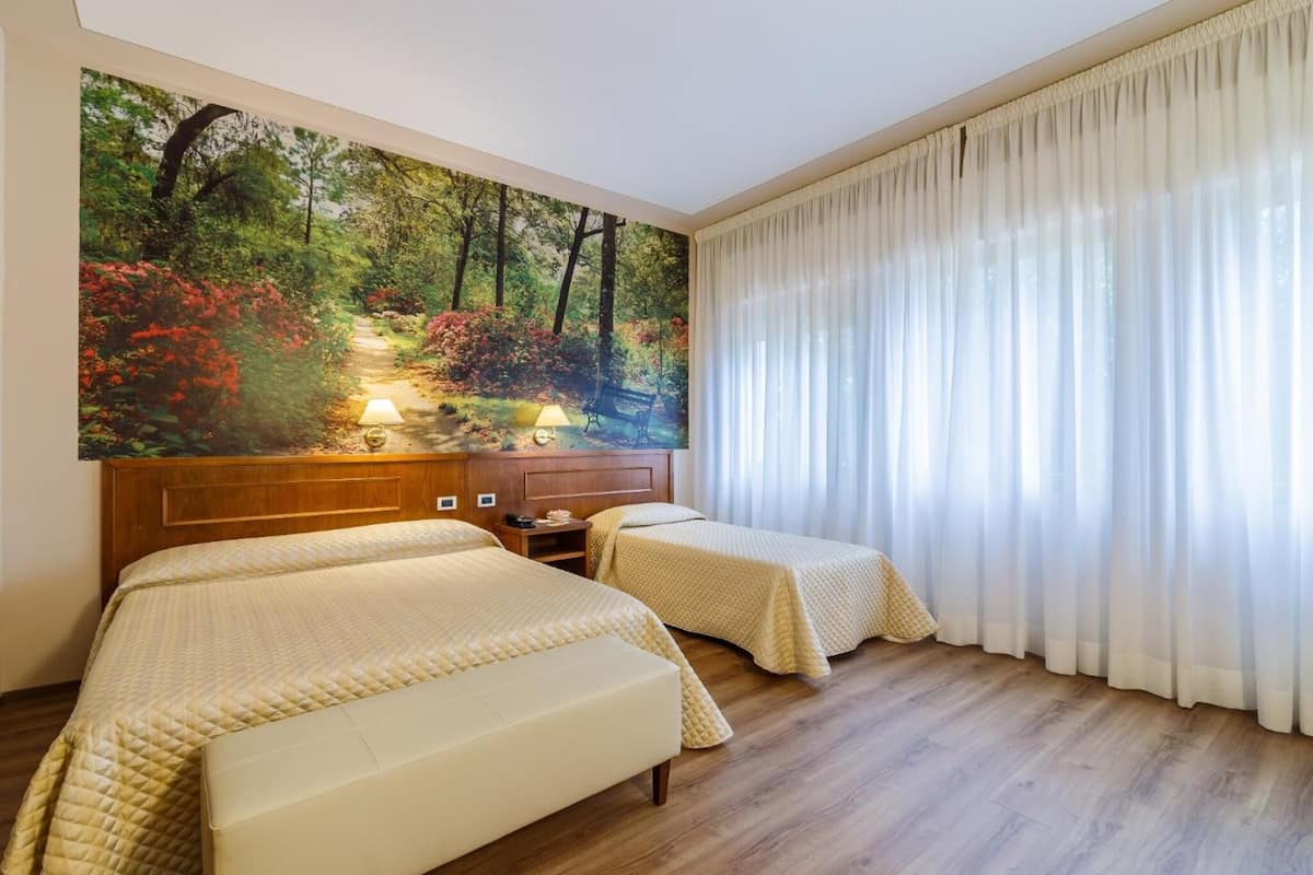 A hotel room with two beds, a large painting of a forest scene, a wooden headboard, and a large window with sheer curtains.