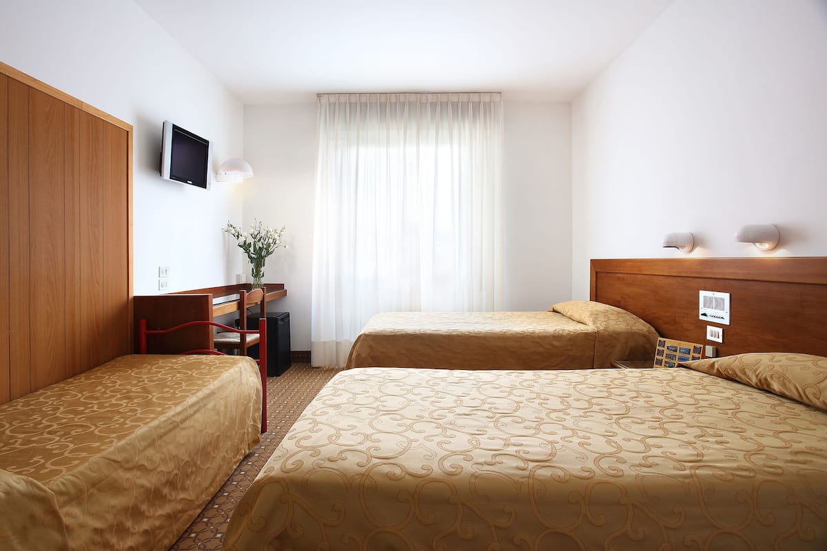 A hotel room with two single beds, a wooden wardrobe, a desk with a chair, a television, and a window with curtains.