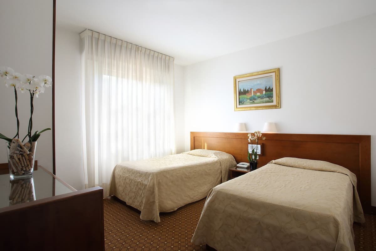 A hotel room with two single beds, a wooden headboard, a nightstand with a lamp, a painting on the wall, and a vase with flowers on a table.
