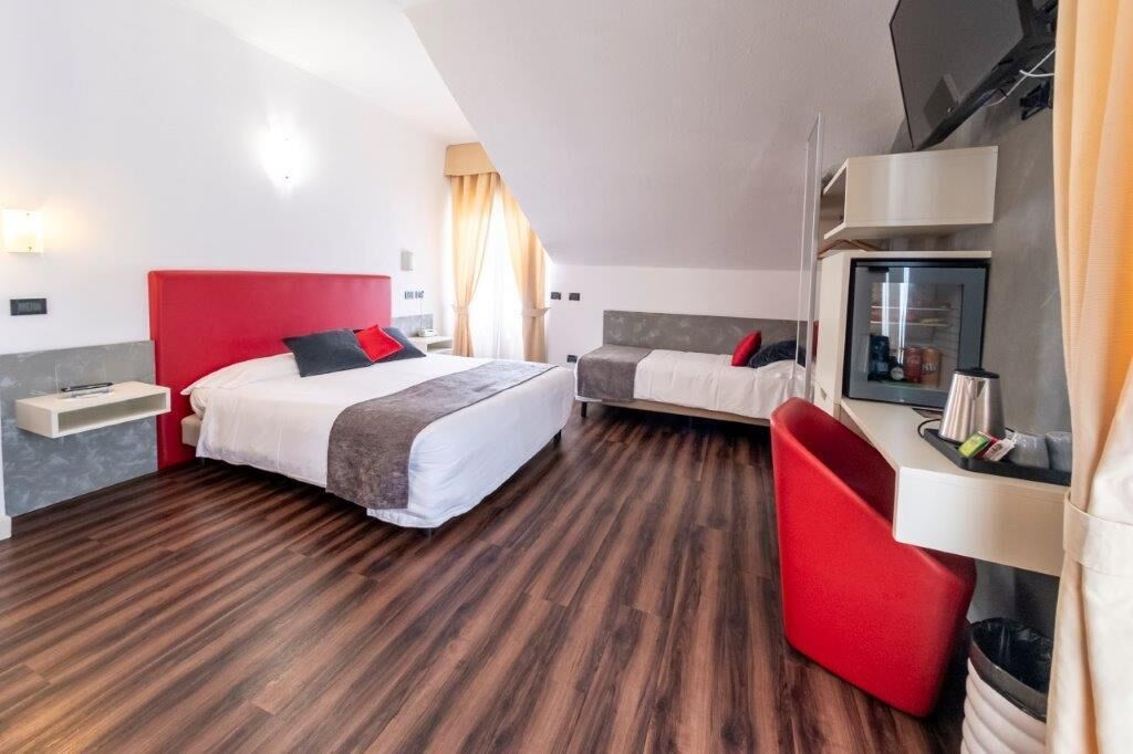 Habitación de hotel con una cama de cabecera roja, otra cama con cabecera gris, un escritorio con una computadora y una silla roja.