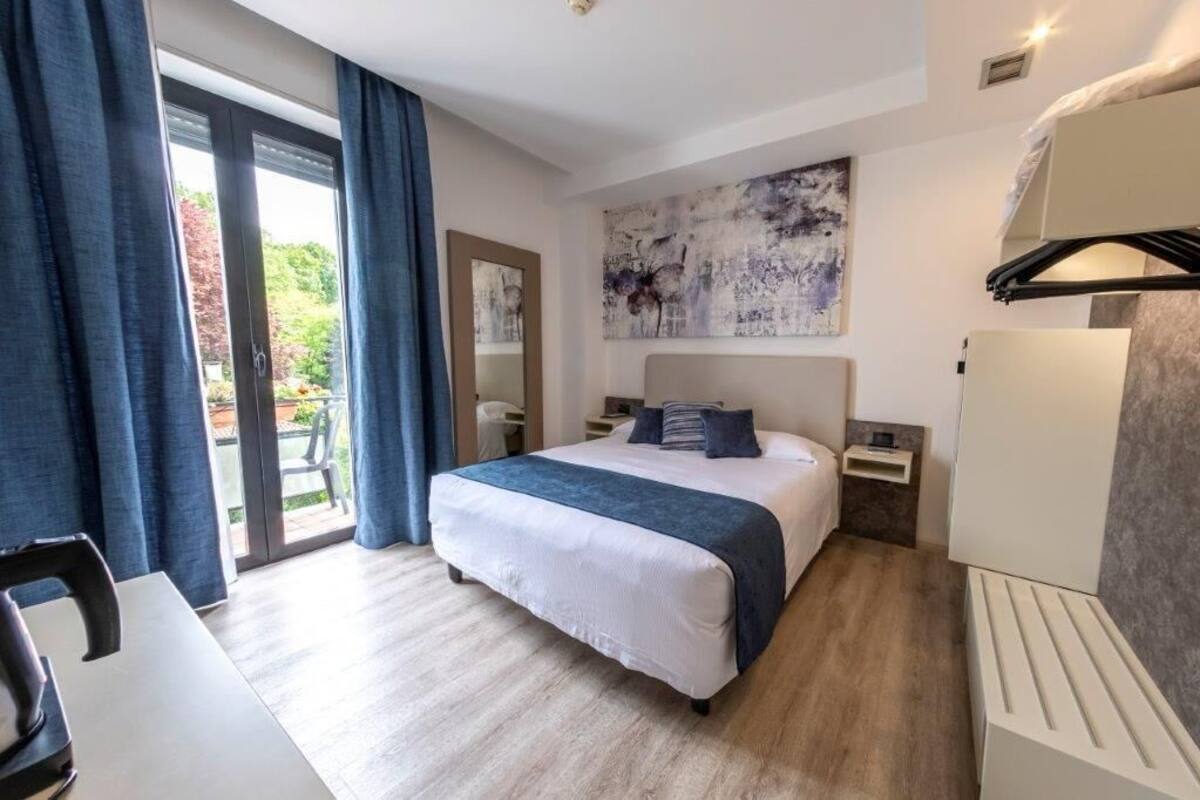 Una habitación de hotel con cama, escritorio, silla, armario y una ventana grande con cortinas.