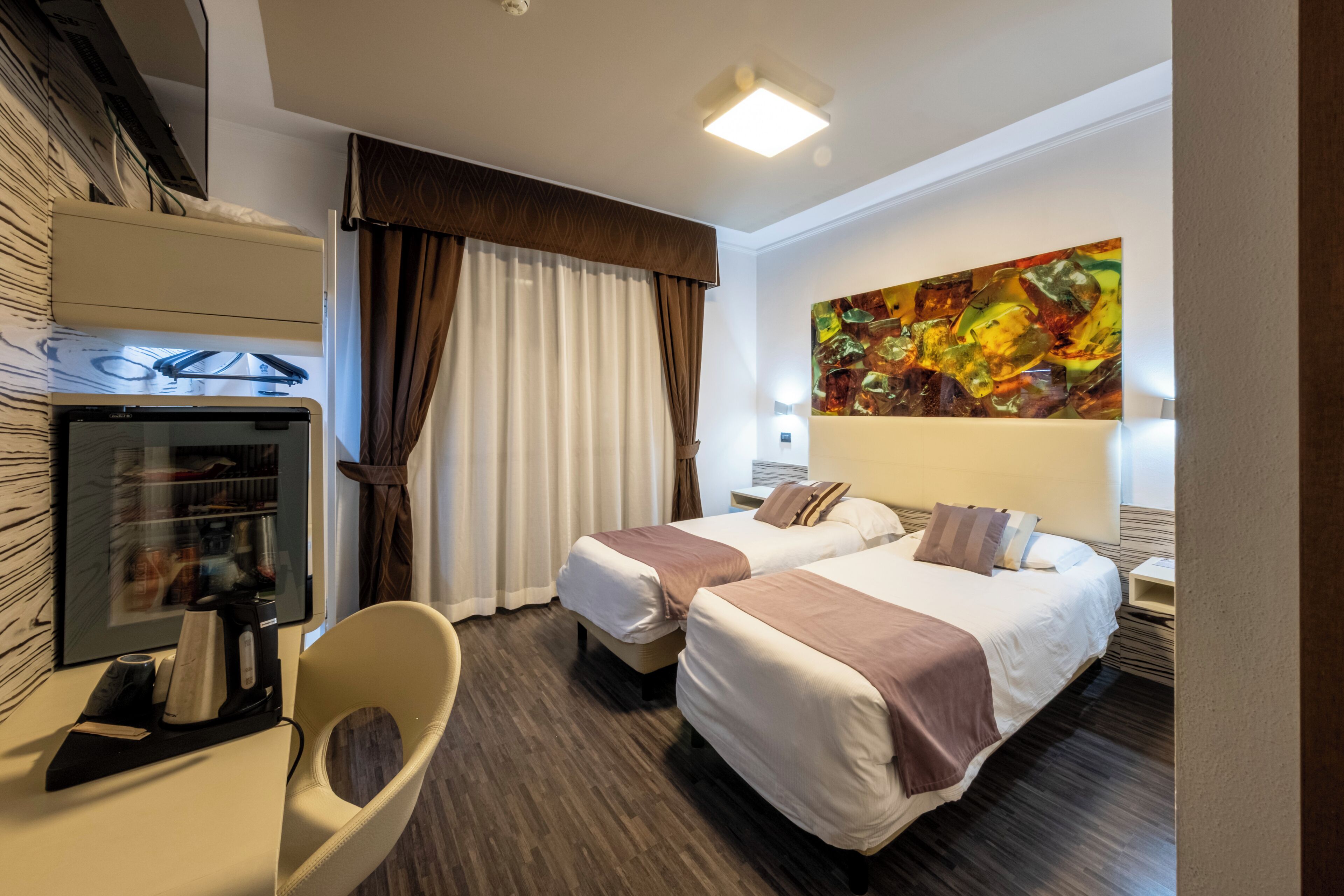A hotel room with two beds, a desk with a coffee maker, a chair, a TV, and a large abstract painting on the wall.