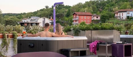 A rooftop terrace with a hot tub, seating area, and a view of a hillside with houses.