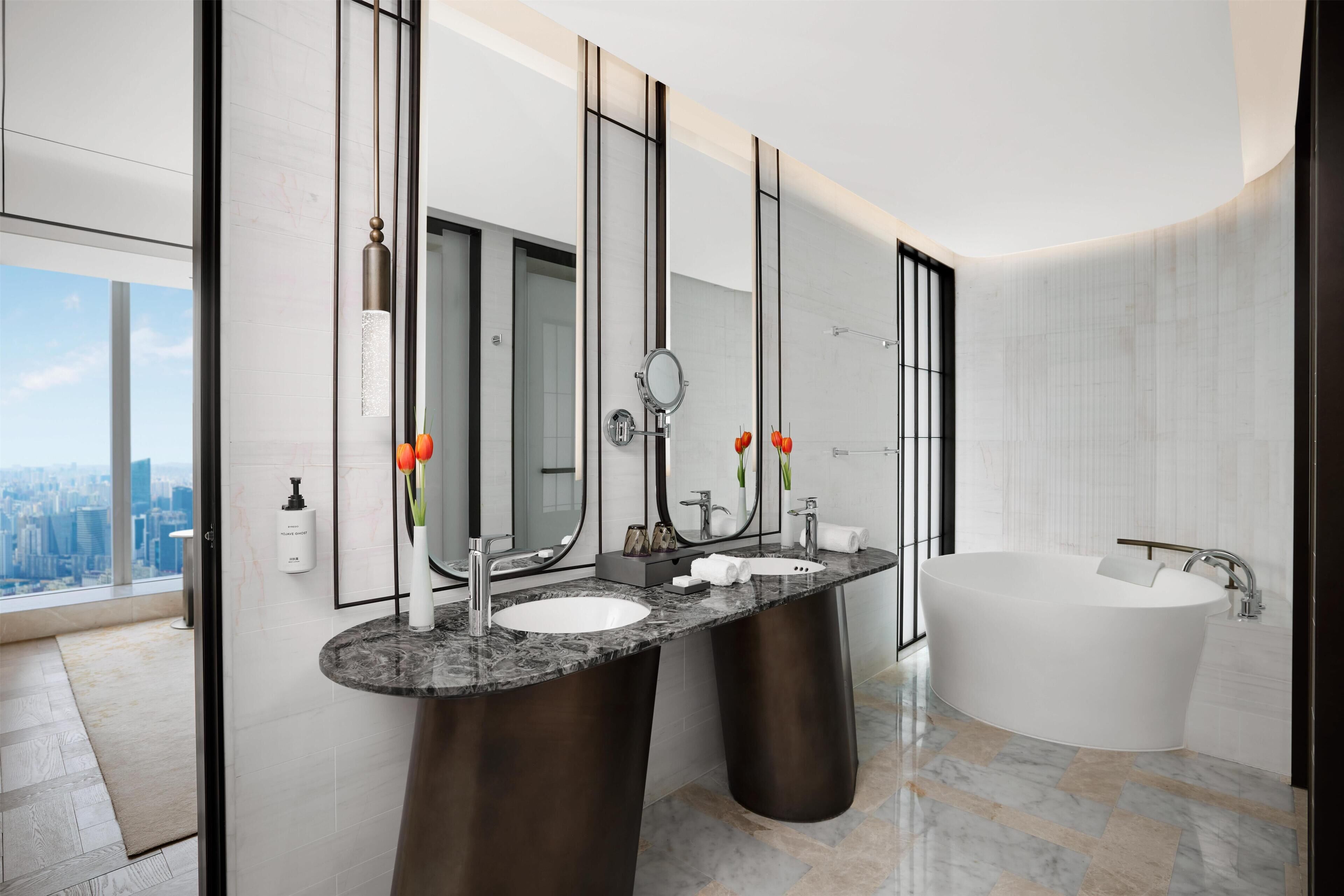 A modern bathroom with dual sinks, a large bathtub, and a city view.