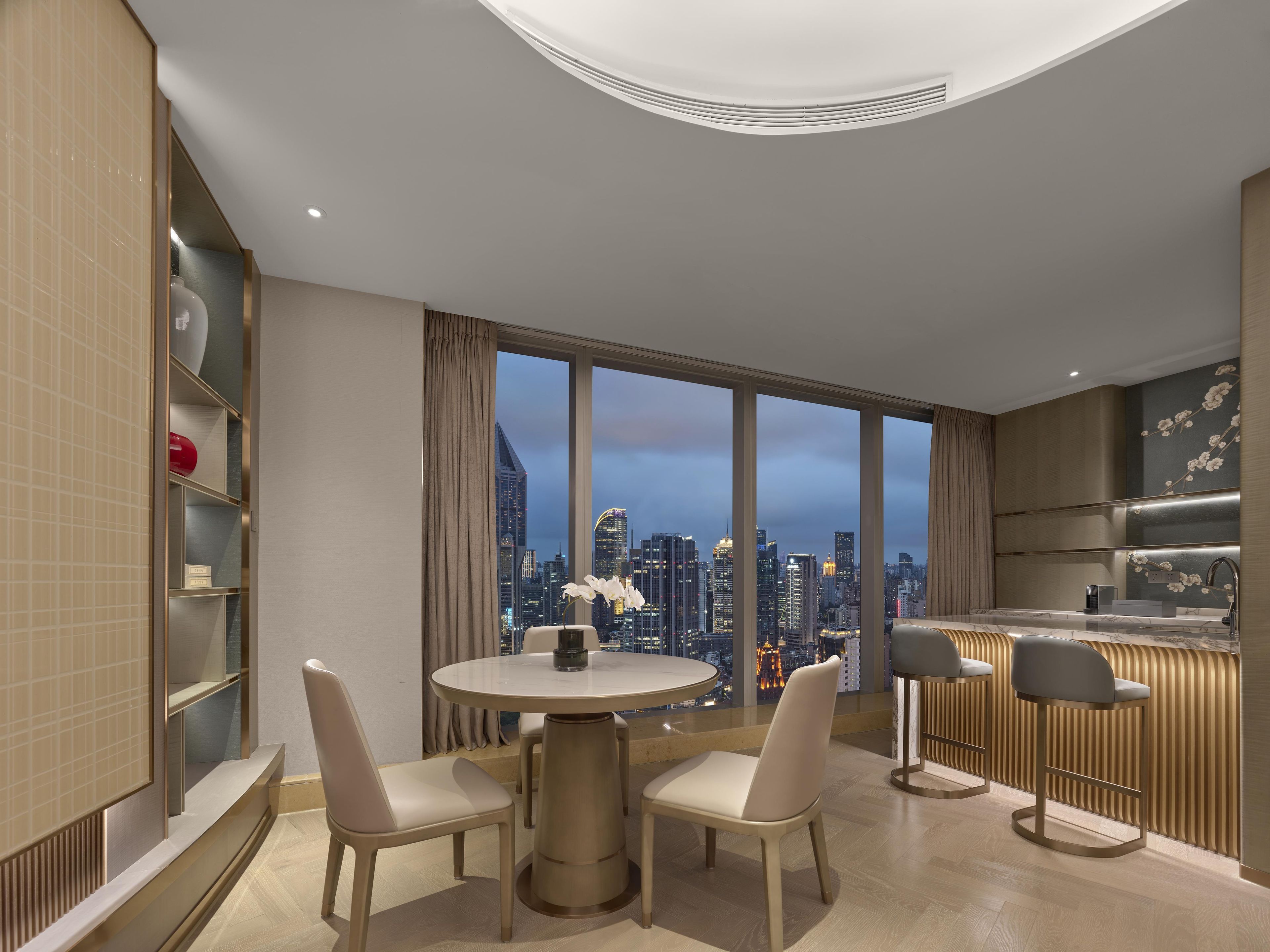 A modern hotel room with a dining area, a kitchen, and a large window with a city view.