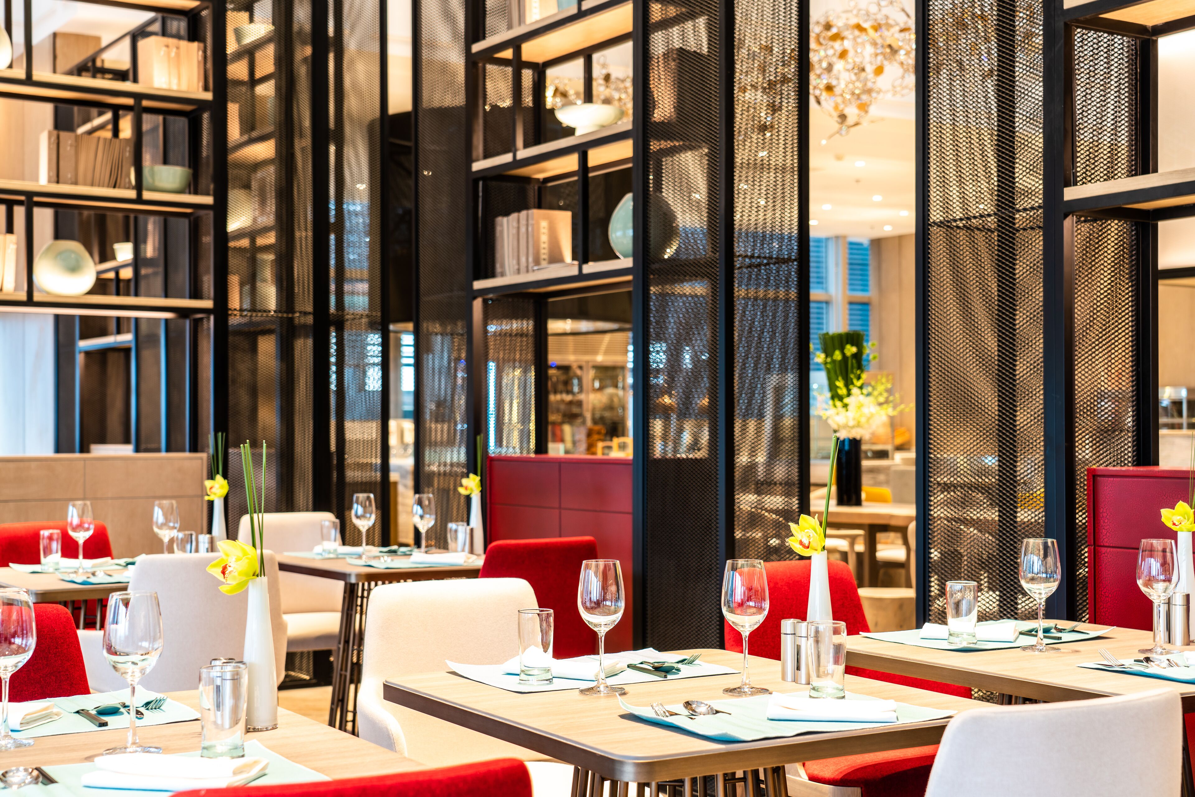 A modern restaurant interior with tables set for dining, red upholstered chairs, and decorative elements like flowers and vases.