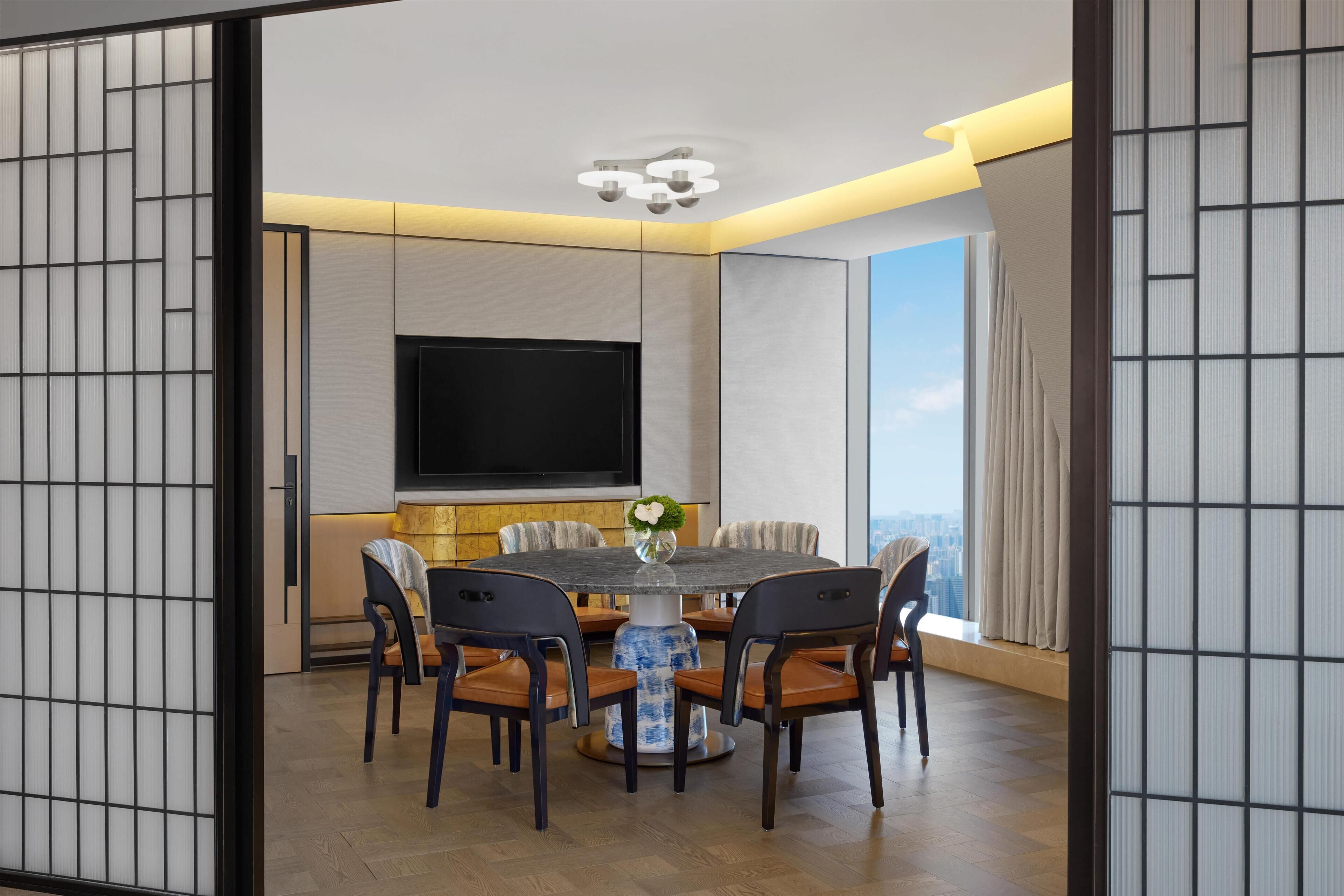 A modern dining area with a round table, chairs, a flat-screen TV, and a city view.