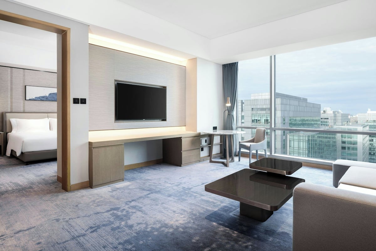 A modern hotel room with a large bed, a desk, a flat-screen TV, a coffee table, and a view of the city.