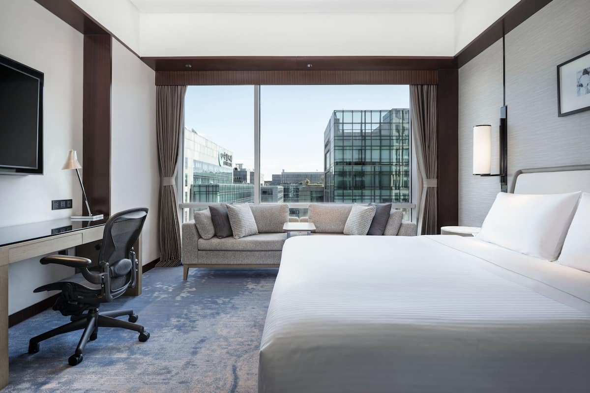 A hotel room with a large bed, a desk with a chair, a sofa, and a large window offering a city view.