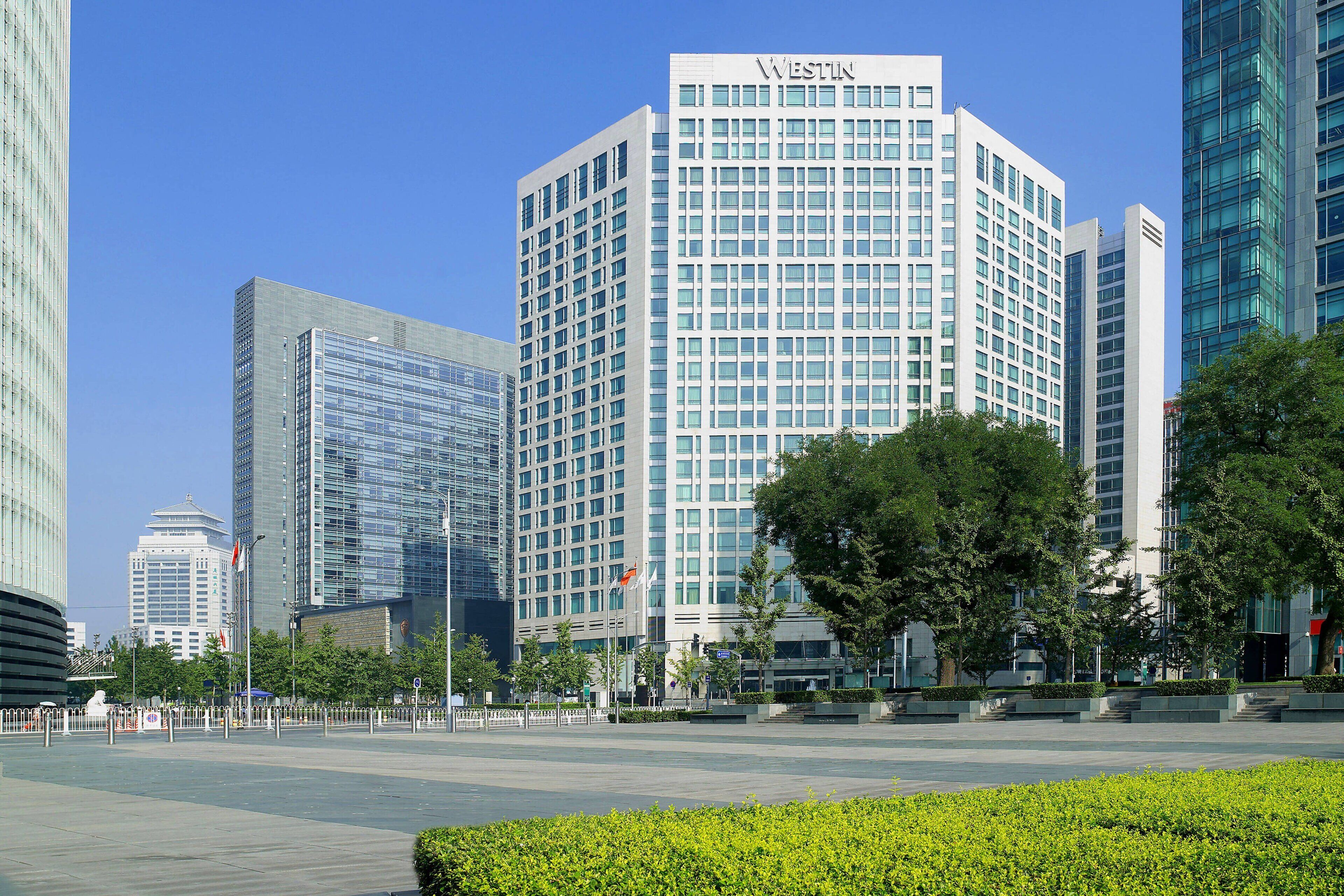 A modern hotel with a prominent 'WESTIN' sign, surrounded by other high-rise buildings and greenery.