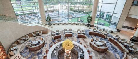 A spacious lobby with a central circular seating area, large windows offering a view of the outdoor courtyard, and a modern staircase on the left.