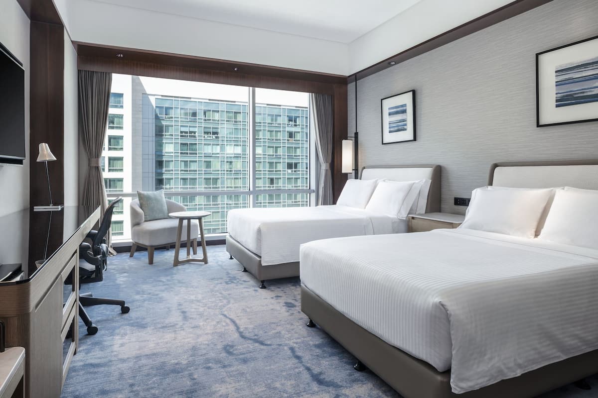 A hotel room with two beds, a desk, a chair, a TV, and a large window with a city view.