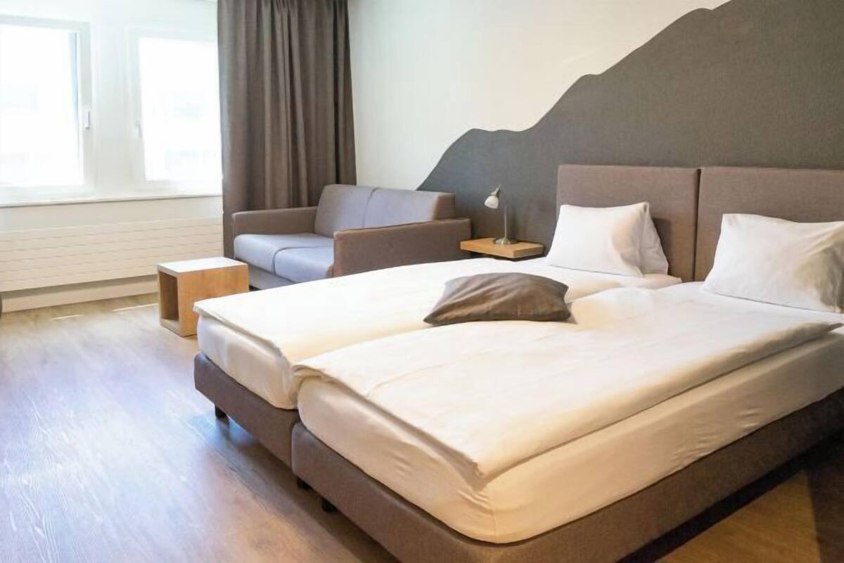 Una habitación de hotel con una cama grande, una silla, una mesita de noche y un ventanal con cortinas.