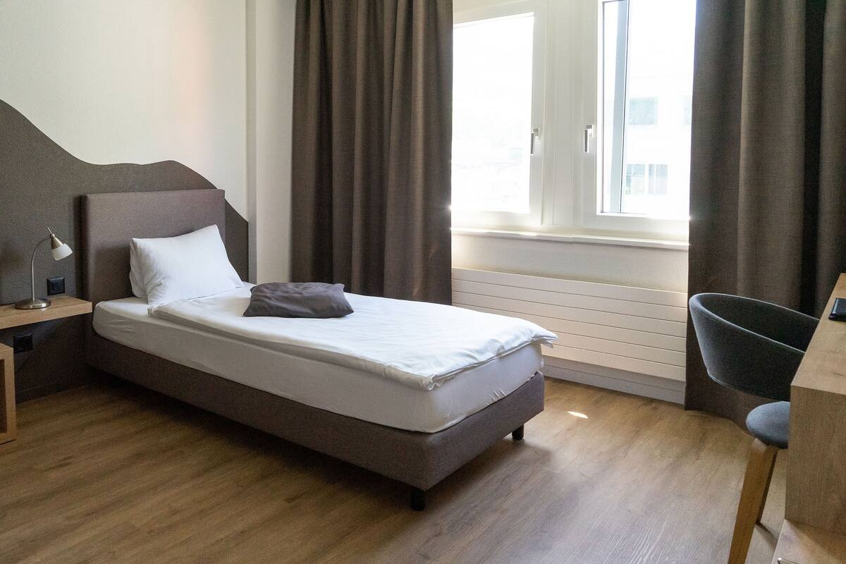 Un dormitorio ordenado con cama, mesita de noche con lámpara, una silla y una ventana con cortinas.