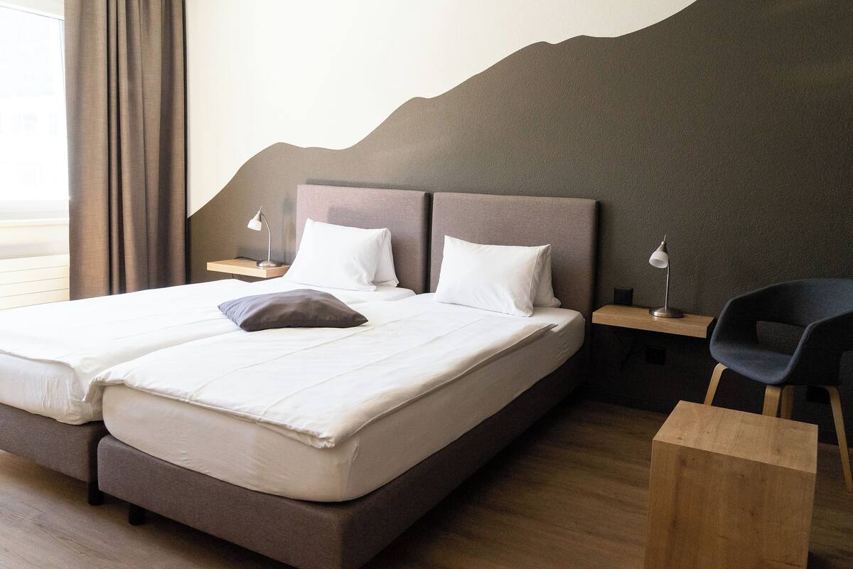 Una cama doble con sábanas blancas, una mesita de noche de madera con una lámpara y una silla moderna.