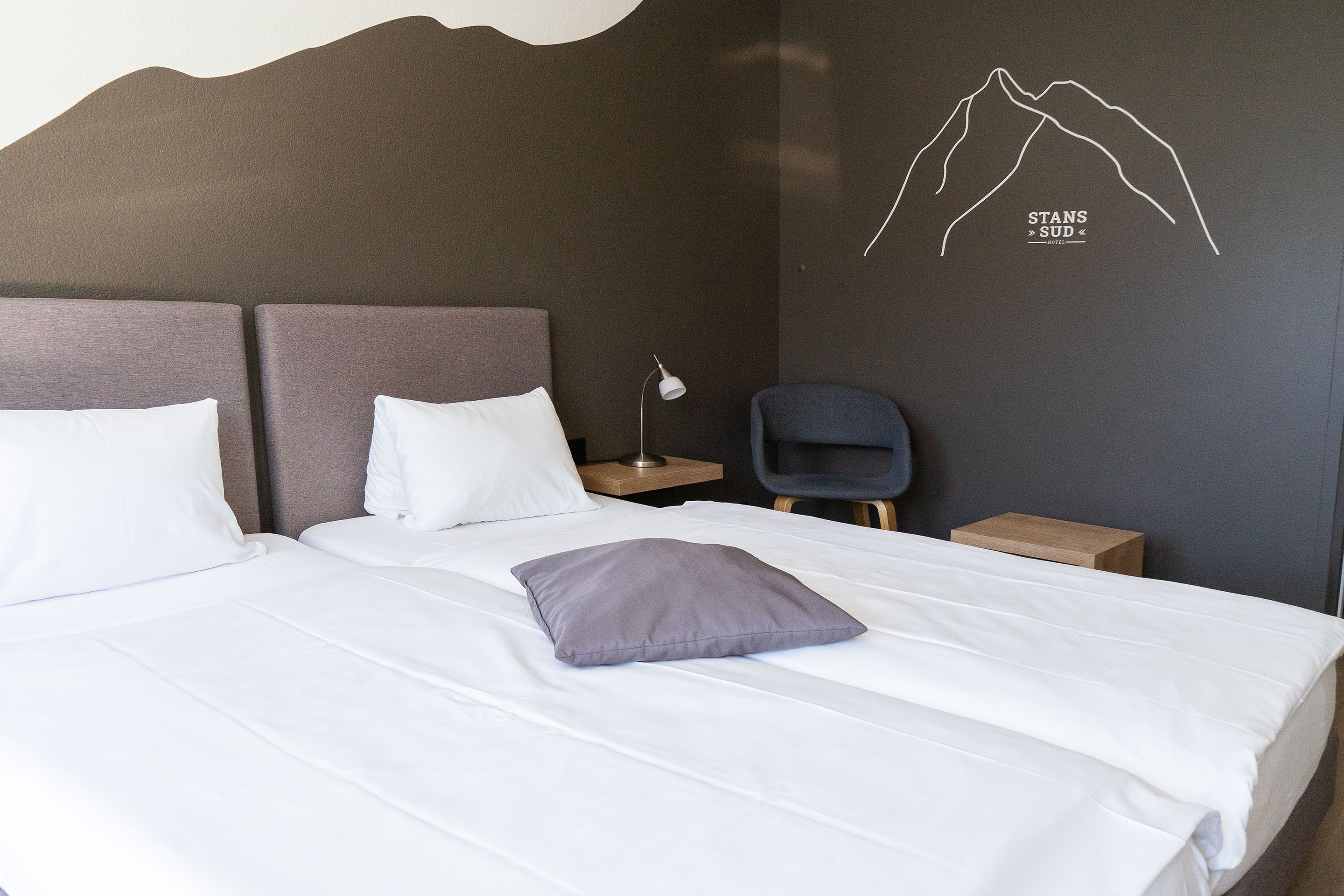 Una cama bien tendida con ropa de cama blanca y gris, una sola almohada gris y un diseño de montaña en la pared.