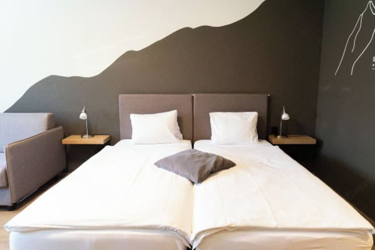 Cama doble con ropa blanca, dos mesitas de noche con lámparas, un sofá gris y una pared oscura con un diseño abstracto en blanco.