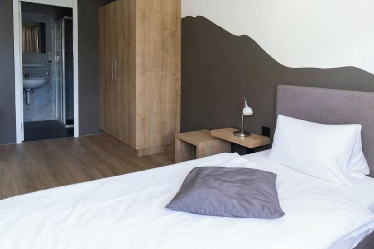 Un dormitorio con cama, mesita de noche, lámpara, armario de madera y baño a la vista a través de una puerta abierta.