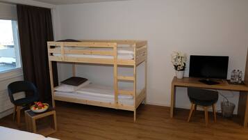A bunk bed with a desk and chair, a television, a small table with a bowl of fruit, and a window with curtains.