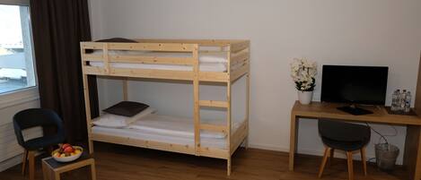 Una cama litera con escritorio y silla, un televisor, una mesita con un frutero, y una ventana con cortinas.