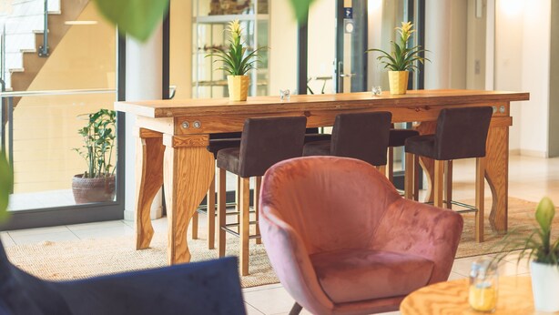 Ein moderner Essbereich mit einem Holztisch, schwarzen Stühlen und einem rosafarbenen Sessel. Auf dem Tisch stehen Blumentöpfe und im Hintergrund eine Treppe.