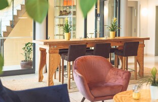 Un área de comedor moderna con una mesa de madera, sillas negras y una silla rosa. Hay plantas en macetas sobre la mesa y una escalera al fondo.