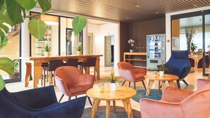 Ein moderner Loungebereich mit Holztischen und -stühlen, einem Automaten und Topfpflanzen.