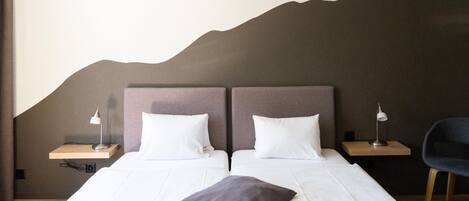 Une chambre d’hôtel avec deux lits, chacun équipé de draps blancs et d’un seul oreiller. On y trouve des tables de chevet avec des lampes, une chaise et un luminaire fixé au mur.