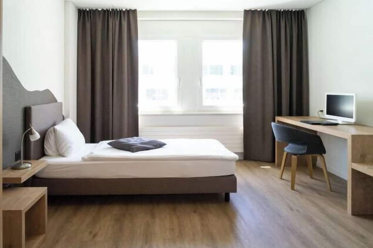 Una habitación de hotel con una cama, un escritorio con computadora y un ventanal con cortinas.
