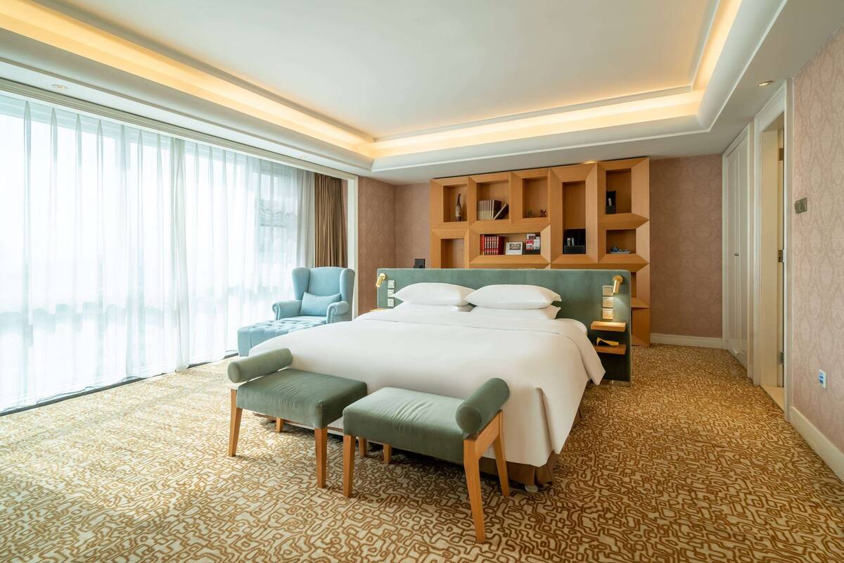 A hotel room with a large bed, a seating area with a bench, a bookshelf, and a view of the city through the window.