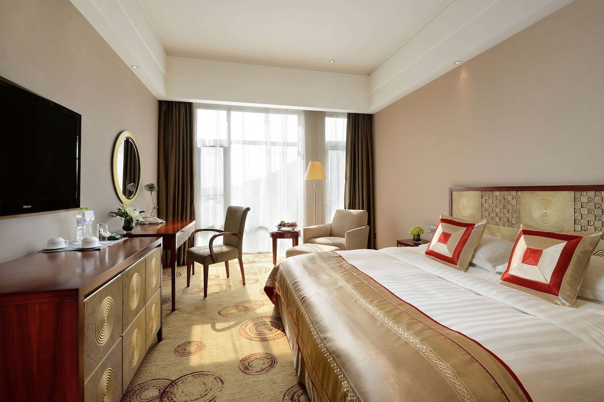 A hotel room with a large bed, a desk with a flat-screen TV, a chair, and a small table.