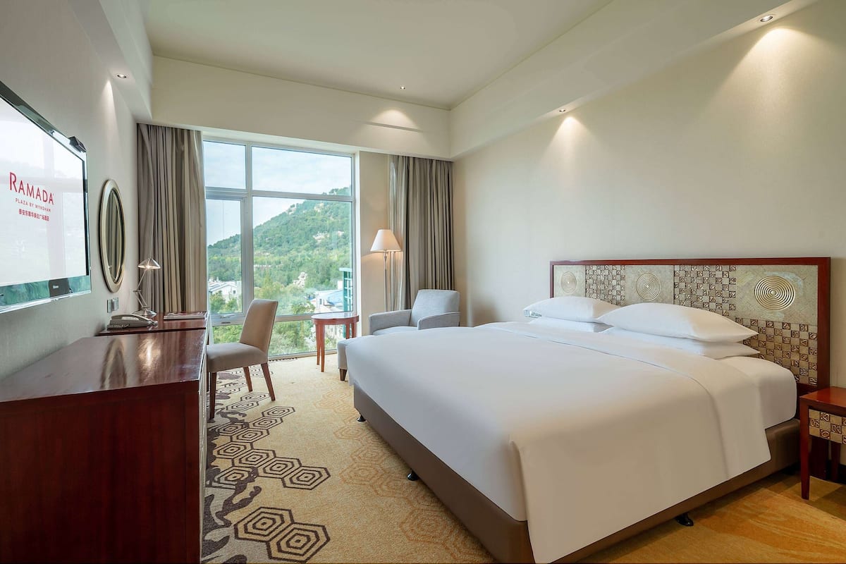 A hotel room with a large bed, a desk, a chair, a lamp, and a view of the outdoors.