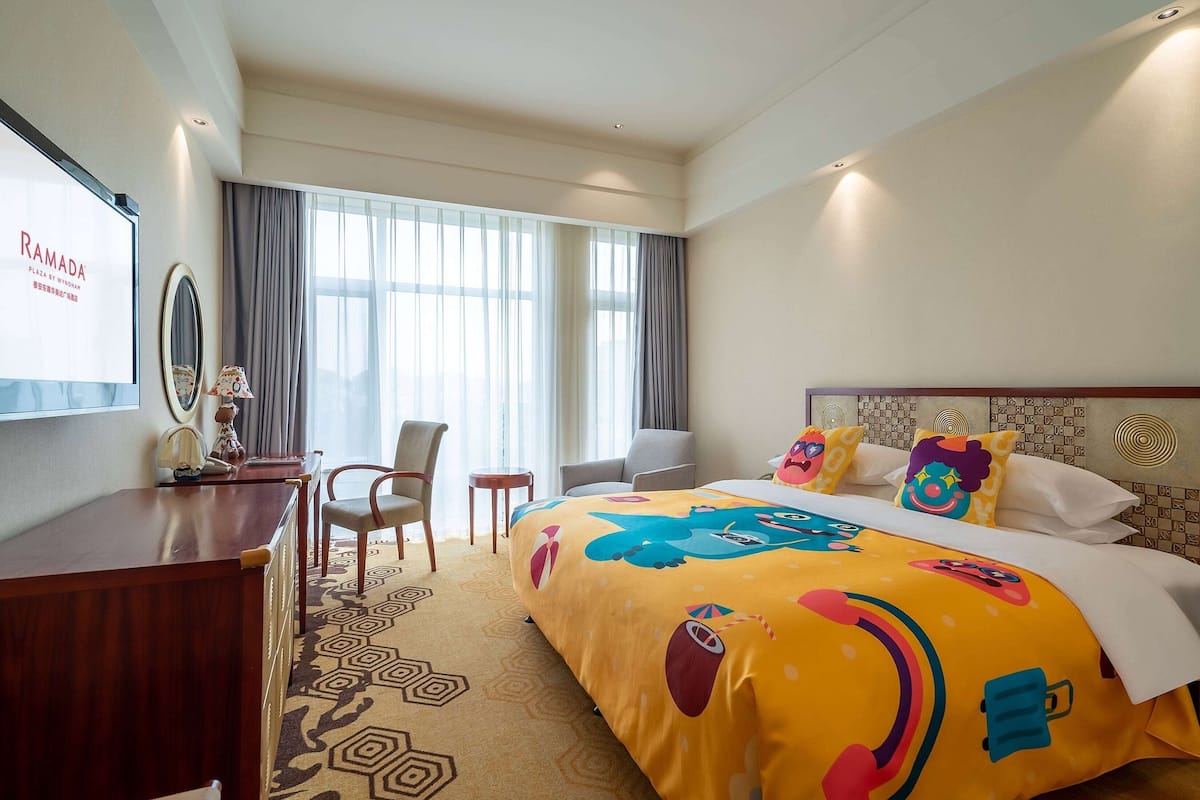 A hotel room with a large bed, a desk, a chair, and a TV screen displaying "RAMADA".