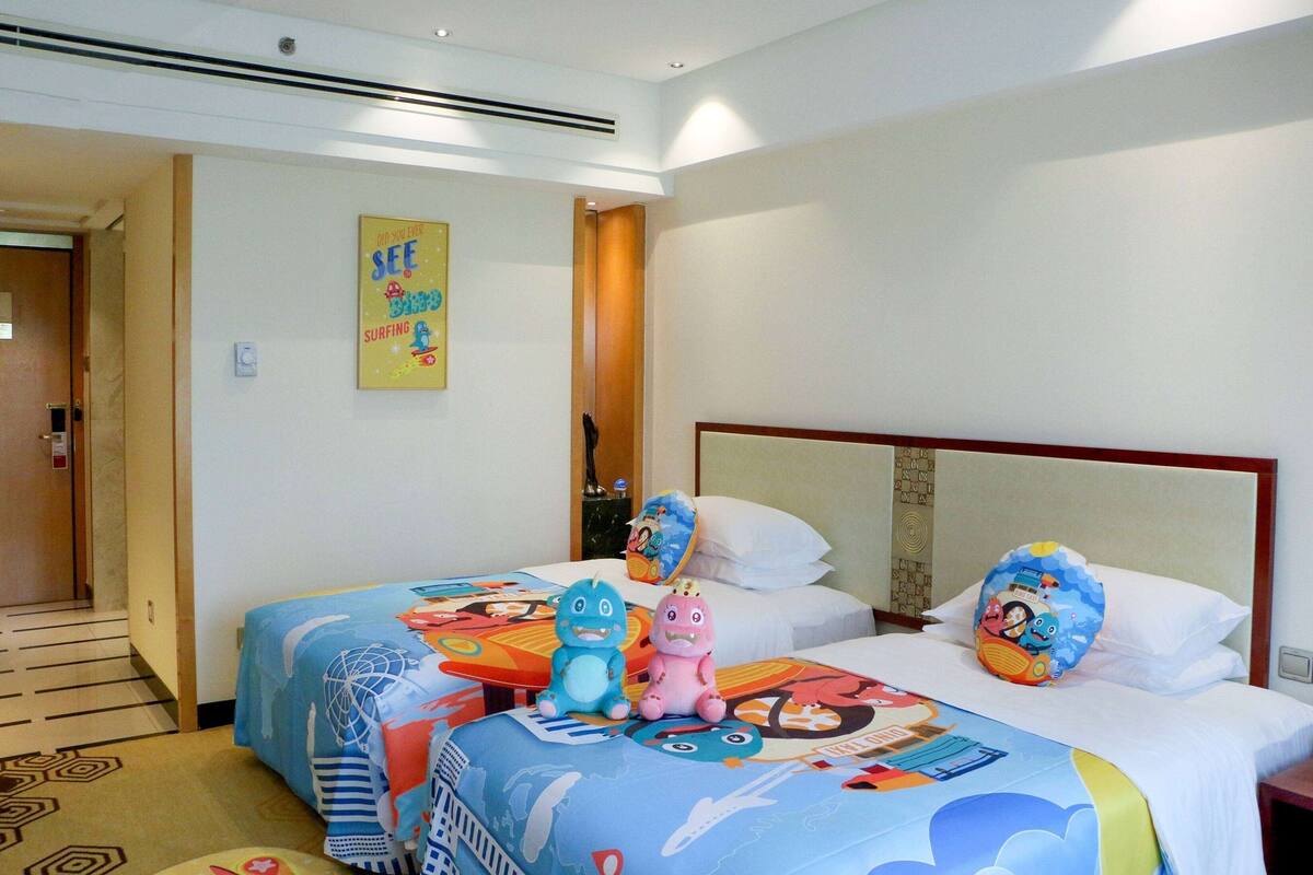 A hotel room with two beds, each with colorful bedding and cartoon character pillows. There is a poster on the wall and a door to the left.