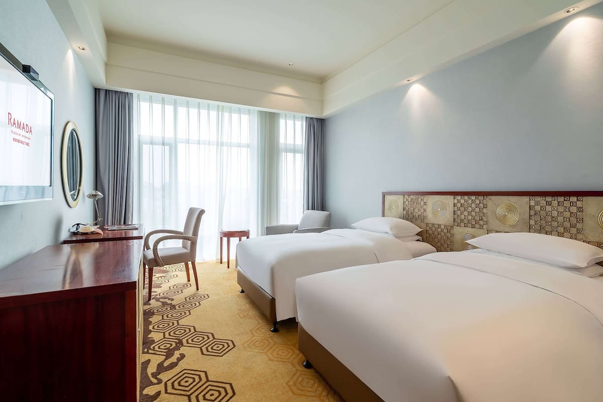 A hotel room with two beds, a desk, a chair, and a large window with curtains.