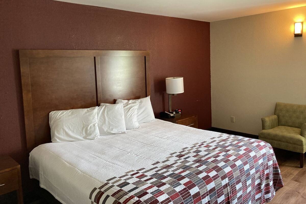 A hotel room with a large bed, a nightstand, a lamp, a chair, and a wall-mounted light.