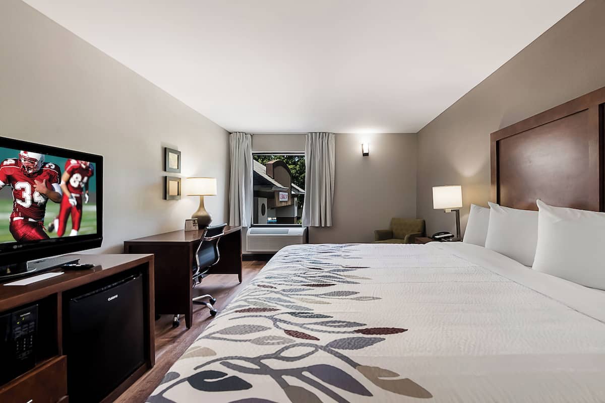 A hotel room with a large bed, a TV on a wooden stand, a desk with a lamp, and a window with curtains.