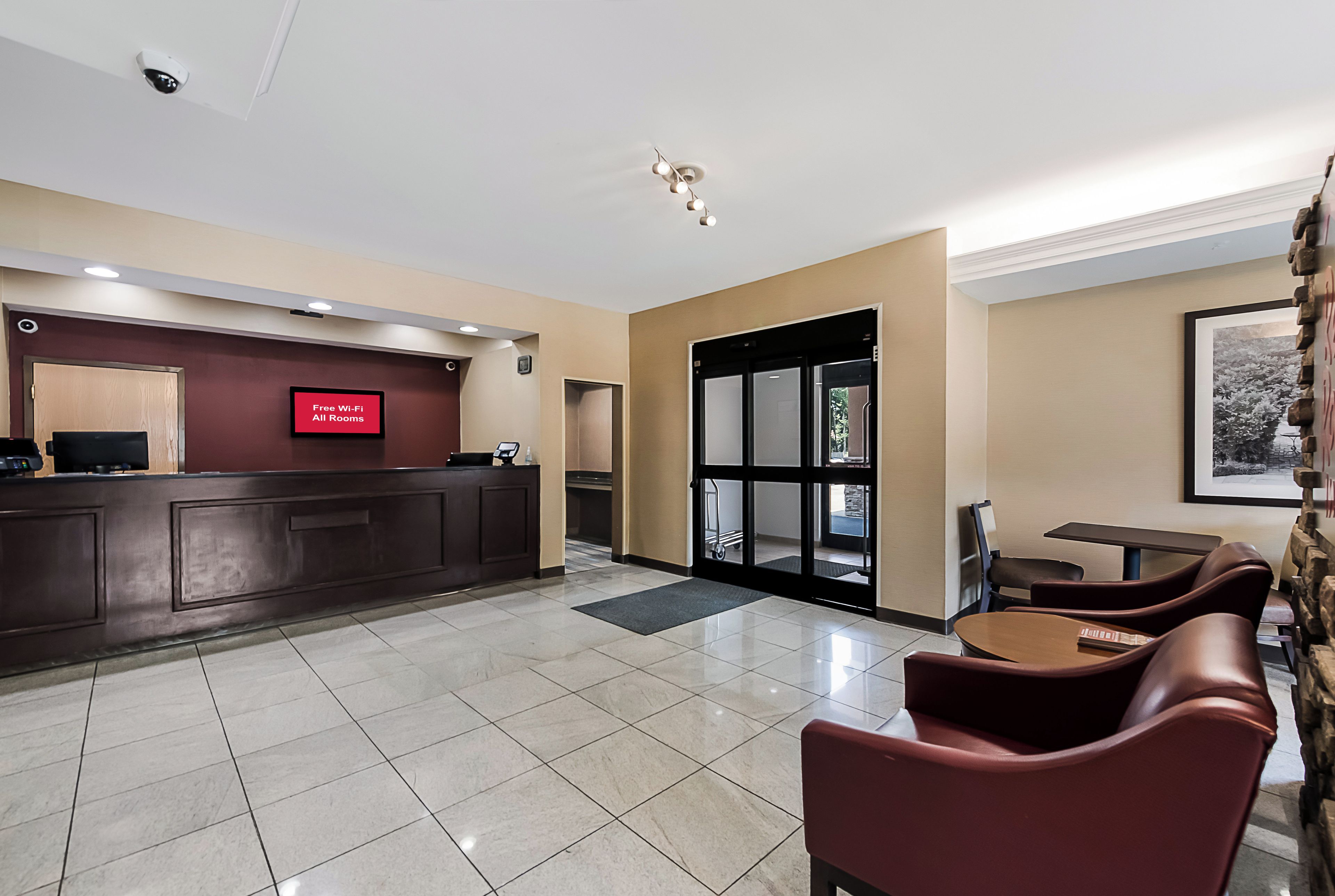 Et hotellets receptionområde med en receptionsdisk, et digitalt skilt der viser 'Gratis Wi-Fi i Alle Værelser', og et siddeområde med stole og et bord.