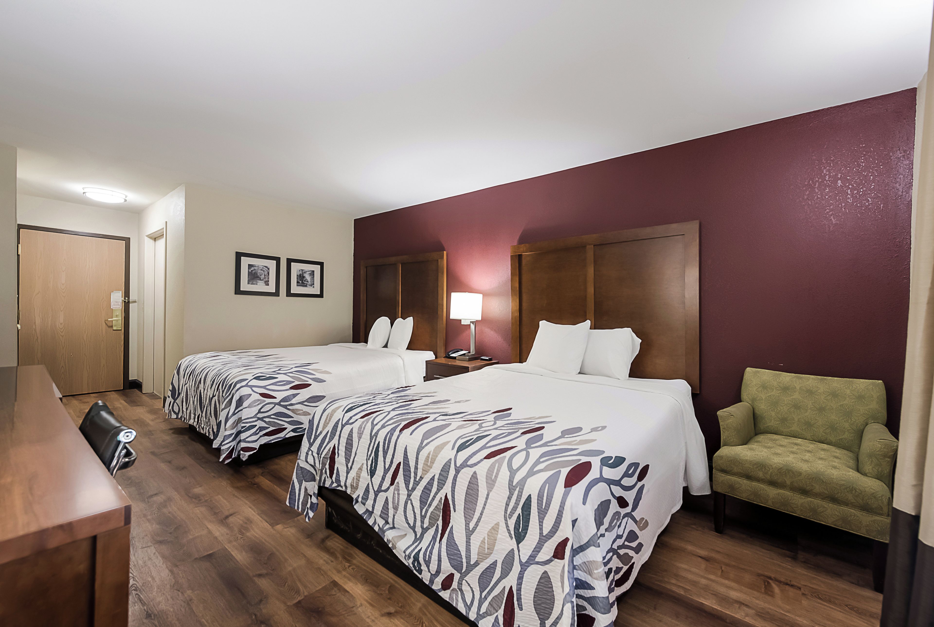 A hotel room with two beds, a desk, a chair, and a nightstand with a lamp.