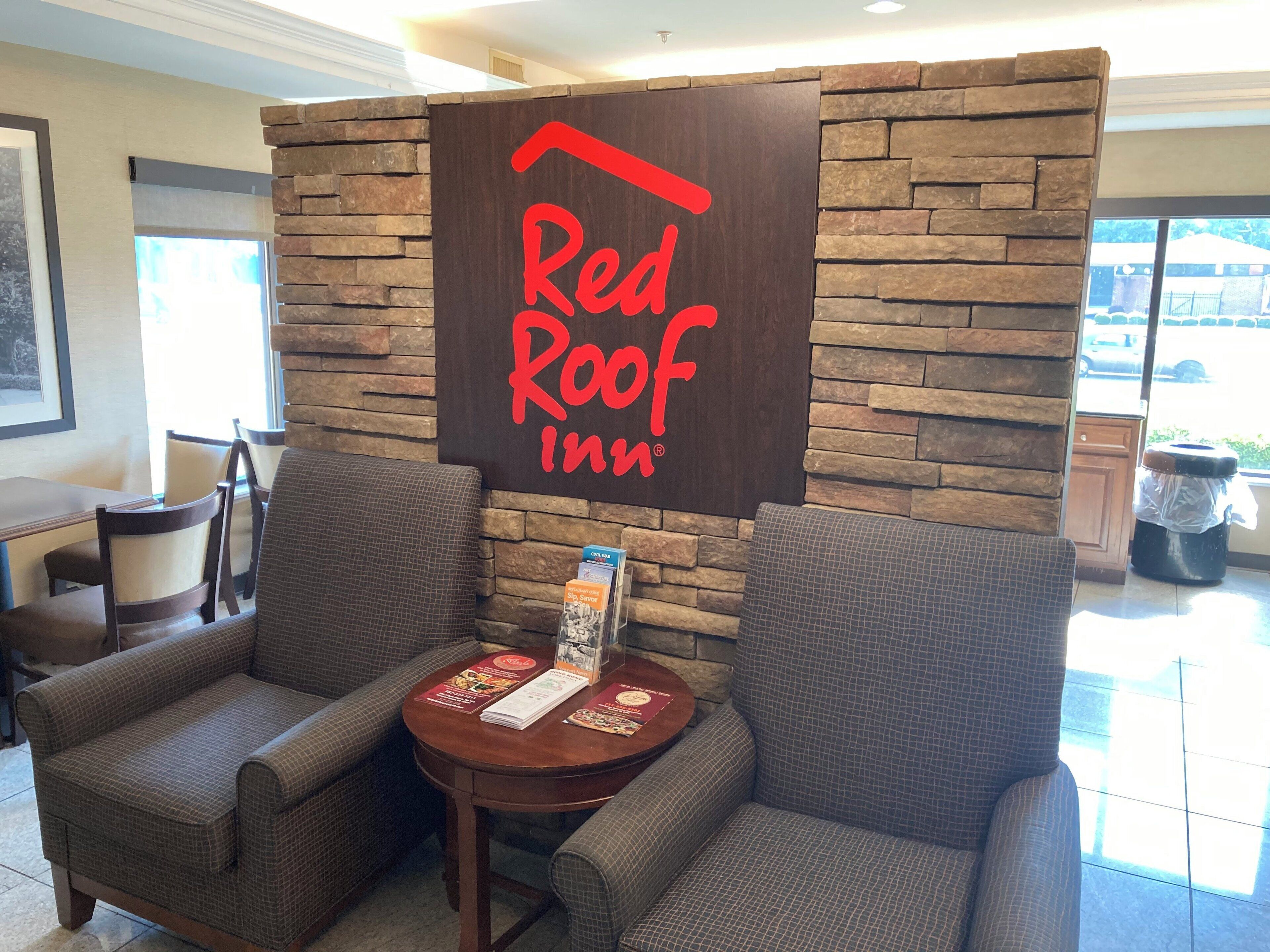 Zona de recepción de Red Roof Inn con un fondo de pared de piedra, dos sillones, una pequeña mesa con un menú y un bote de basura.