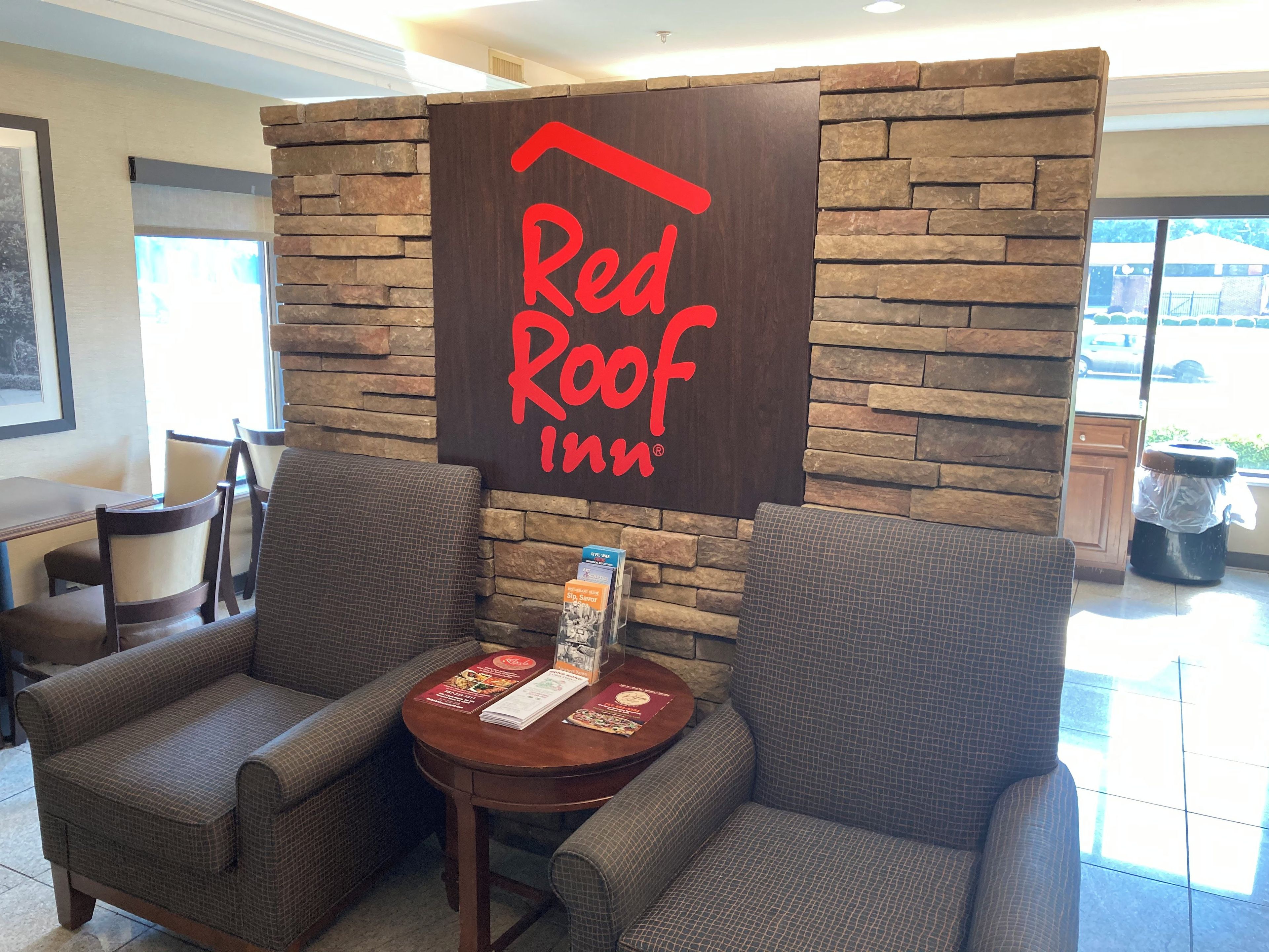 A Red Roof Inn reception area with a stone wall backdrop, two armchairs, a small table with brochures, and a trash can.