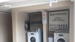 Zona de lavandería con dos lavadoras, un dispensador de jabón con varios productos y un cartel de prohibido fumar.
